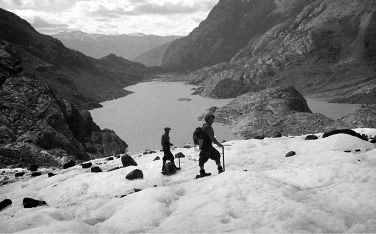 Two explorers with ice axes walk across a snow-covered glacier, with a lake and mountains in the background