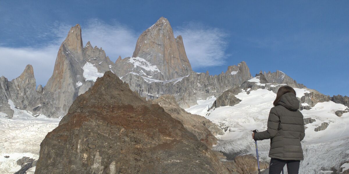 Hiker with trekking poles seen from behind on the crest of the moraine at Laguna De los Tres, facing the granite walls of Fitz Roy and its glaciers