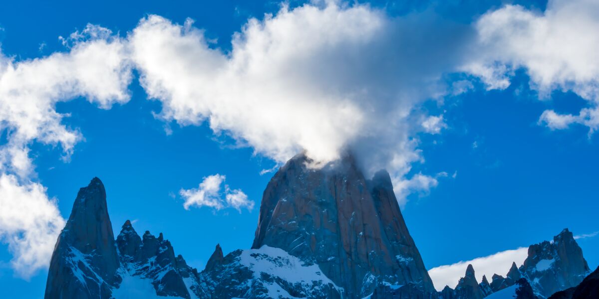 The granite summit of Fitz Roy with clouds streaming off its peak, beside other snowy spires, under a blue sky with sunlit clouds