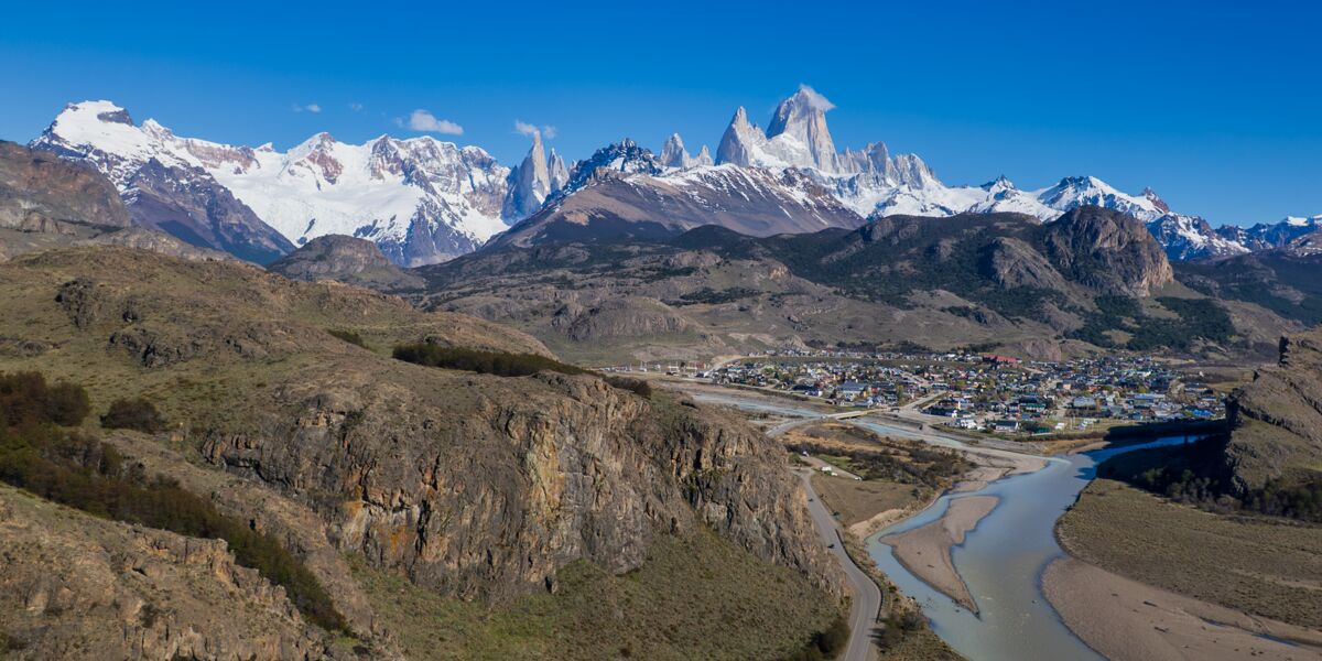 Aerial view of El Chaltén beside the De las Vueltas River, with snow-capped Cerro Torre and Fitz Roy behind it under a blue sky