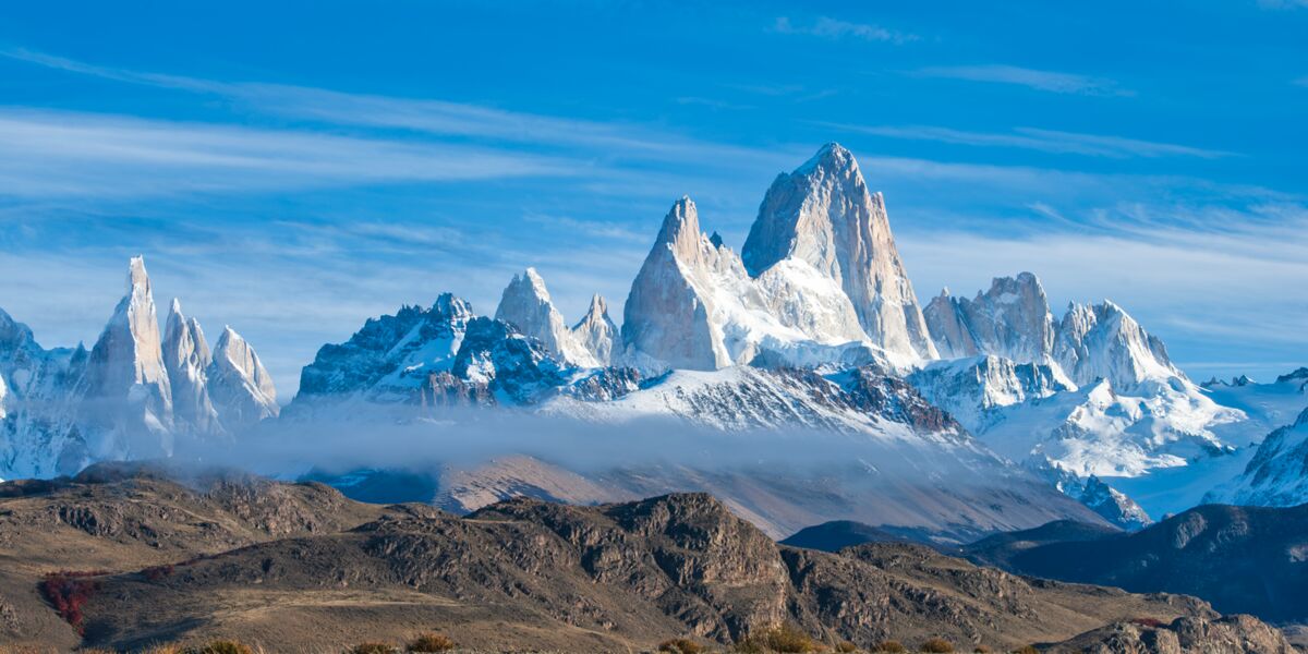 The Cerro Torre range and the Fitz Roy massif covered in snow under a blue sky, with a band of low clouds over the valley