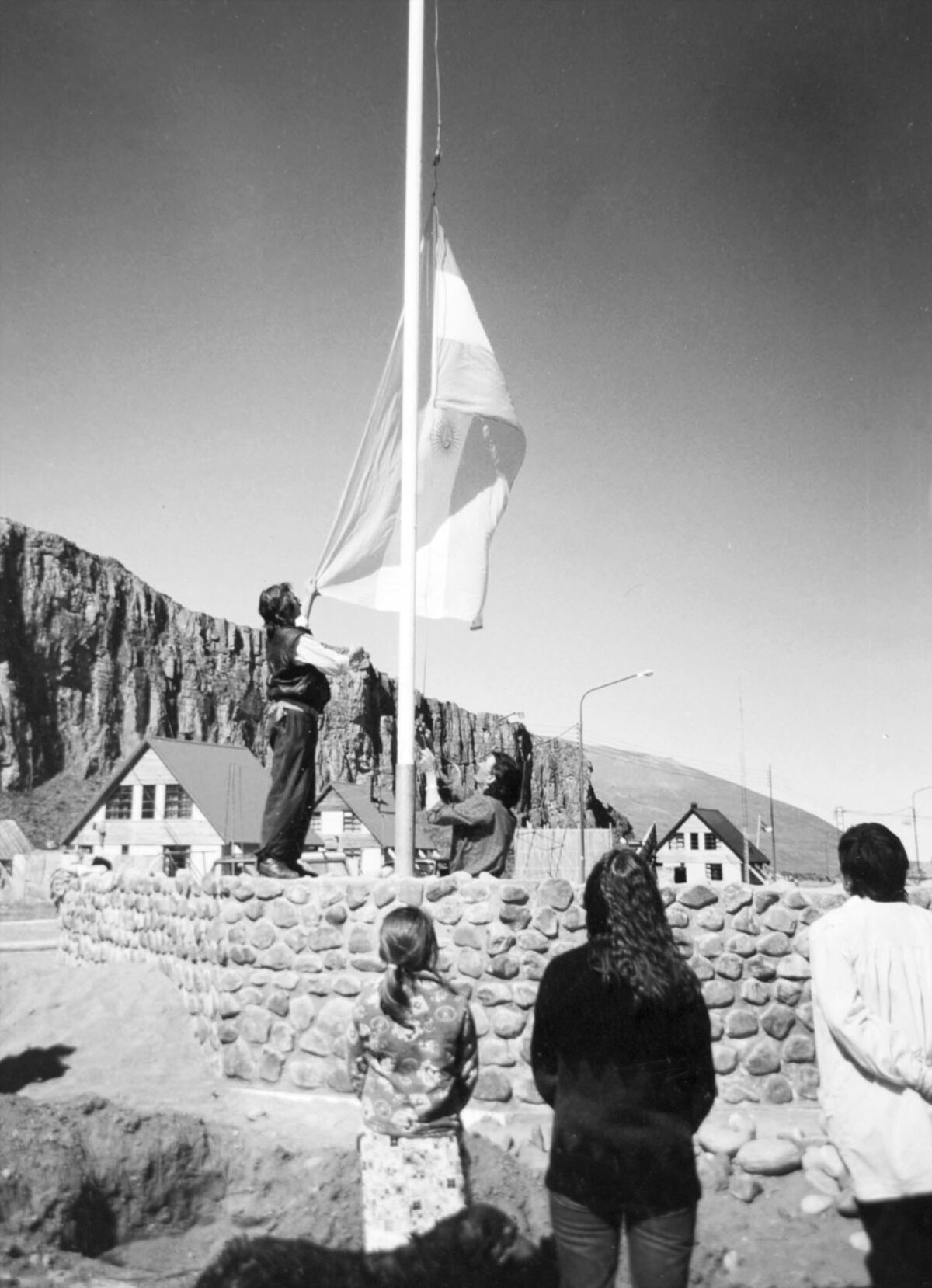 Residents of El Chaltén raise the Argentine flag on the town flagpole, with a rock wall and gabled houses in the background