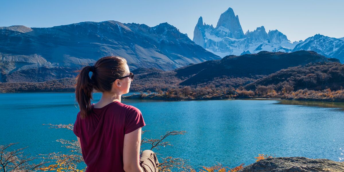 Caminante con anteojos de sol frente a la Laguna Capri, con el cerro Fitz Roy despejado al fondo y lengas con colores de otoño