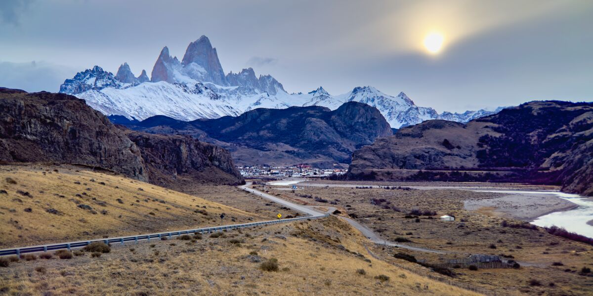 The road descends toward El Chaltén along the Río de las Vueltas, with snow-capped Mount Fitz Roy behind the town
