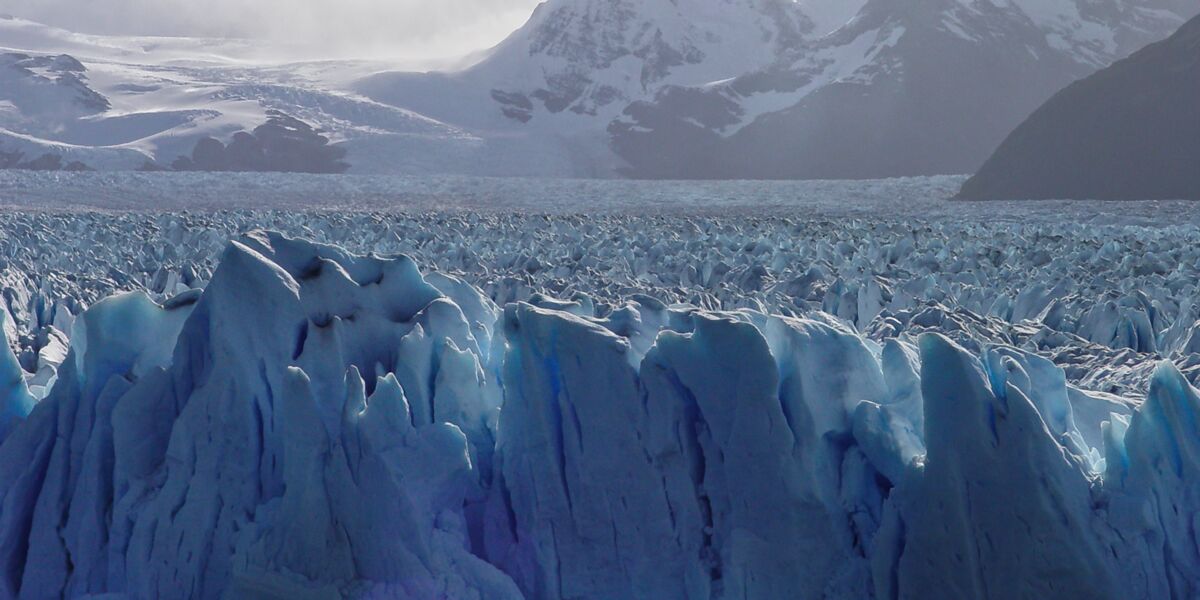 Surface of the Perito Moreno Glacier covered in crevasses and seracs of blue ice, with snow-capped mountains behind